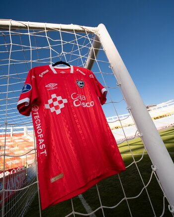 Camiseta futbolística roja del club Cienciano colgada de una percha en la red de la arquería dentro de un estadio de fútbol
