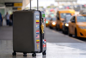 Una maleta gris decorada con pegatinas de banderas de países y un llavero del trofeo Mundial, en un aeropuerto con taxis amarillos al fondo