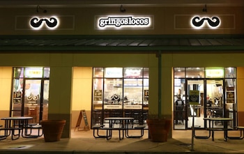 Vista frontal de un restaurante Gringos Locos con letrero luminoso, logo de bigote, ventanas, mesas, bancos, jardineras y un cartel tipo sándwich