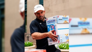 Hombre con gorra blanca Wilson Sporting Goods y polo negro con logo NB carga tres cajas apiladas de galletas y galletitas saladas