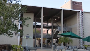 Edificio con ladrillo claro, estructura de concreto, escaleras y balcones exteriores. Leíble: "NATURAL & ENVIRONMENTAL SCIENCES". Hay sombrillas verdes y bancos