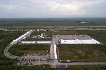 Fotografía aérea de un extenso complejo de carpas blancas y edificios modulares en un terreno plano y verde. Se observan aparcamientos, caminos y una pista de aeropuerto