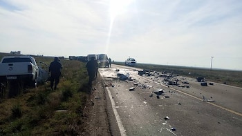 Carretera asfaltada con escombros, una camioneta blanca con puerta abierta, dos personas, un vehículo policial y varios camiones al fondo, cielo claro