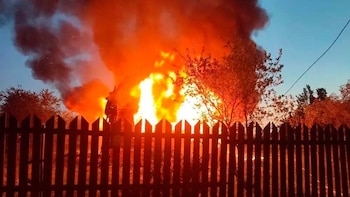 Imagen de un gran incendio emitiendo llamas brillantes y humo oscuro, visible detrás de una valla de madera. Árboles silueteados se ven a los lados, y el cielo azul oscuro es visible arriba