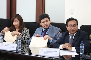 Diputados sentados en una mesa curva con micrófonos, documentos, botellas de agua e identificadores de nombres en una sala de reuniones