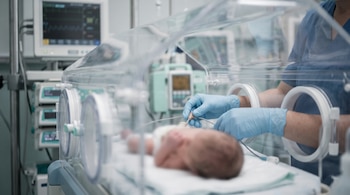 Un bebé dentro de una incubadora médica y unas manos con guantes que colocan un sensor frente a varios monitores clínicos.