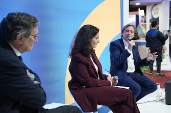 Tres ponentes, dos hombres y una mujer, sentados en un escenario azul y amarillo. Un hombre habla por micrófono. La pantalla muestra el título del evento. Público en primer plano