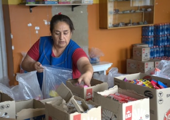 Mujer con delantal azul y camiseta roja empaca productos de cajas en bolsas de plástico; cajas de alimentos y botellas de agua apiladas al fondo