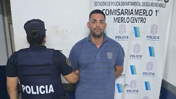 Un hombre con barba y polo azul a rayas es retenido por una policía de espalda. Detrás hay un cartel de la Comisaría Merlo 1° con logos de Policía