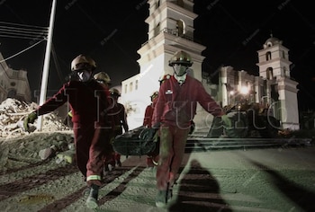 Cinco socorristas con casco, mascarilla y uniforme rojo cargan una camilla sobre escombros de edificios destruidos en Pisco durante la noche