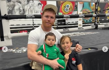 Canelo Álvarez, un hombre de tez clara y barba rojiza, sonríe a la cámara mientras carga a una bebé vestida de verde. A su derecha, una niña pequeña también sonríe, en un gimnasio de boxeo con un ring