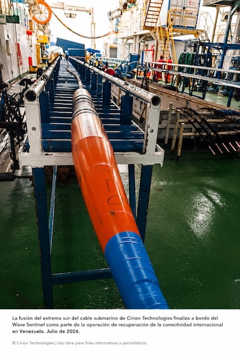 Un cable submarino grueso de color naranja y azul sobre una estructura metálica en la cubierta de un barco, con maquinaria y escaleras al fondo