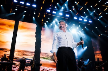 Ricardo Montaner, con camisa blanca y pantalón negro, sonríe en un escenario oscuro con luces brillantes y pantallas que muestran un atardecer. Sostiene un micrófono