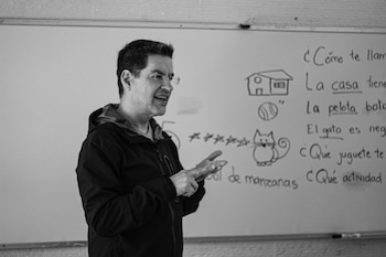 Hombre con lentes y chaqueta oscura gesticula frente a un pizarrón blanco con dibujos de una casa, una pelota, un gato y texto escrito en español