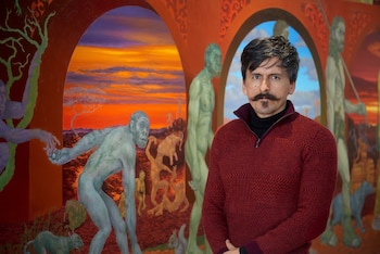 Un hombre con bigote y suéter rojo posa delante de una pintura con arcos y figuras humanas antiguas