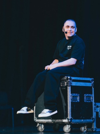Lucho Miranda, un hombre calvo con una camiseta negra y audífono, sentado en una caja de equipo de sonido negra con ruedas sobre un escenario oscuro