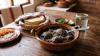 Cazuela de barro con mole prieto y ajonjolí, plato con arroz y frijoles, cesta con tortillas y vaso con agua sobre una mesa de madera.
