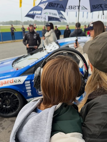 En un circuito, mujeres y niños con auriculares disfrutan del evento. Se ve un coche de carreras azul y una mujer con abrigo rojo en el pit lane