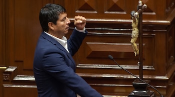 Un hombre con traje azul y camisa blanca levanta el puño derecho frente a un micrófono y un crucifijo, con paneles de madera de fondo.