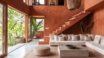Un espacioso salón con paredes terracota, suelo de concreto, sofá claro, mesa de centro de concreto y un gran ventanal que da a un jardín tropical.