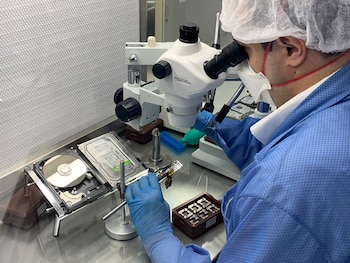 Un hombre con mascarilla, bata y guantes examina las piezas de un disco duro abierto mediante un microscopio sobre una mesa de trabajo