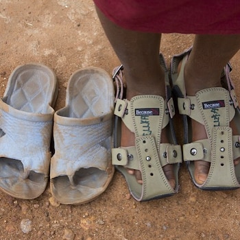 Unos pies calzan sandalias ajustables con correas junto a un par de chanclas desgastadas sobre la tierra
