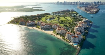 Vista aérea de Fisher Island con edificios, campo de golf, playas de arena blanca y aguas turquesas; el horizonte de Miami se ve en la distancia