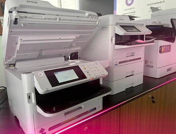 Varias impresoras Epson blancas, algunas multifunción, sobre una superficie oscura con luz de neón rosa. Una impresora tiene la tapa superior abierta y la pantalla encendida