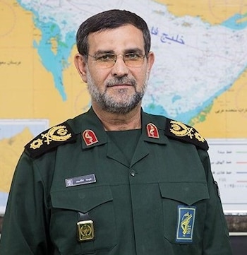Retrato de Alireza Tangsiri, un hombre con gafas, barba, vestido con un uniforme militar verde oliva, frente a un mapa geopolítico