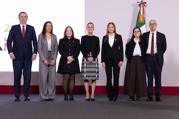 Siete personas vestidas con ropa formal posan de pie en fila sobre un escenario con la bandera de México al fondo
