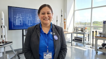 Retrato de Patricia Ortiz, ingeniera aeroespacial, sonriendo en un entorno de laboratorio con maquetas de cohetes y un monitor que muestra planos de naves.