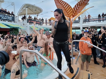 Una cantante con tatuajes y ropa oscura actúa con un micrófono en la cubierta de un crucero, rodeada por una multitud y personas en un jacuzzi en primer plano