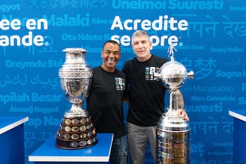 Dos hombres sonrientes, uno de ellos es Cafu, de pie junto a dos trofeos plateados, con un fondo azul que contiene texto en varios idiomas