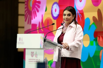Una mujer en un atril con micrófonos y el logotipo del Gobierno de la Ciudad de México frente a un panel decorativo de colores