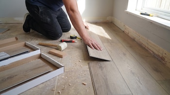 Un hombre arrodillado ajusta una tabla de piso de madera junto a herramientas como un mazo, un cúter y una cinta métrica.