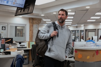 El actor Noah Wyle lleva una sudadera gris y una mochila al hombro mientras camina por la recepción de un centro médico