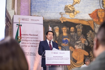 Hombre en traje oscuro con corbata habla en un podio con micrófonos, frente a banners blancos con logos del Gobierno de México. Al fondo, un mural