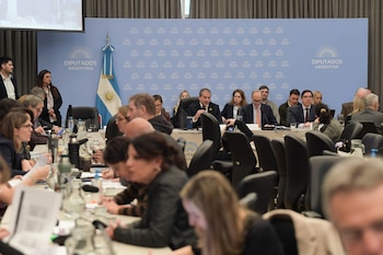 Personas sentadas en mesas de trabajo dentro de una sala con un panel institucional y una bandera de Argentina