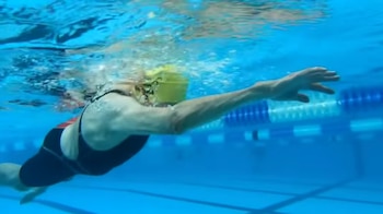 Nadadora con gorro amarillo y traje de baño oscuro bajo el agua. Extiende un brazo en brazada dentro de una piscina con líneas de carril