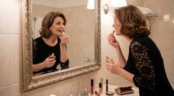 Mujer de mediana edad con cabello castaño, vestida de negro, se aplica pintalabios rojo mientras sostiene un frasco de perfume frente a un espejo ornamentado en un baño.