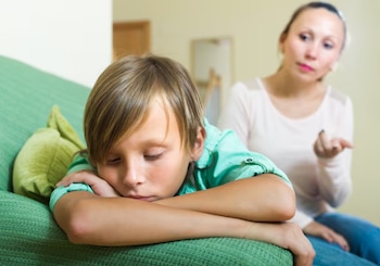 Niño recostado en un sofá verde con almohada. Mujer adulta, borrosa, en segundo plano, con la mano abierta