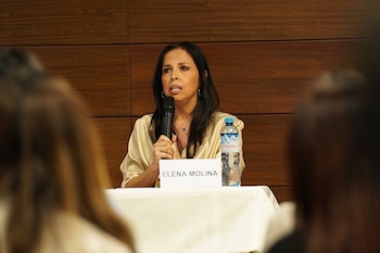 Una mujer sentada detrás de una tarjeta con el nombre Elena Molina sostiene un micrófono frente a personas desenfocadas