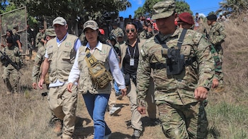 Mujer con chaleco y gorra avanza por sendero de tierra. La acompañan hombres en uniforme militar de camuflaje y otros con chalecos caqui