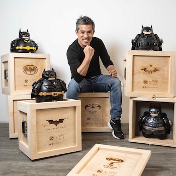 Marcelo Wong sonríe junto a esculturas negras de Batman con formas redondeadas colocadas sobre cajas de madera