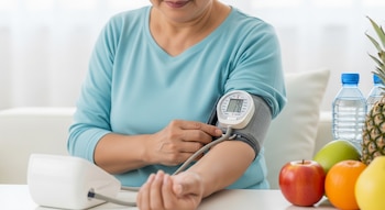 Vista cercana de una persona adulta midiéndose la presión arterial en el brazo con un tensiómetro digital. En la mesa hay varias frutas y botellas de agua.