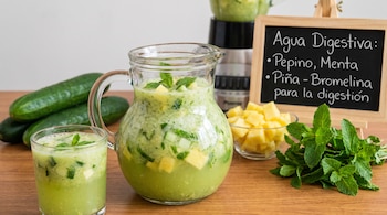 Jarro con bebida verde y menta, vaso con la misma bebida, pepinos, piña en cubos, hojas de menta, licuadora y cartel con texto en español.