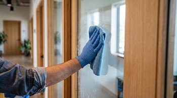 Una mano con guante azul pasa un paño de microfibra azul sobre una puerta de vidrio con marco de madera.