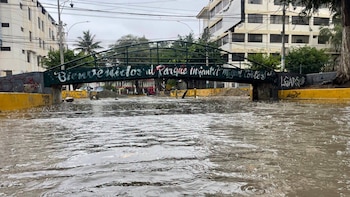 Una vía inundada por agua de lluvia bajo un puente con la inscripción Parque Infantil Miguel Cortés, entre edificios urbanos