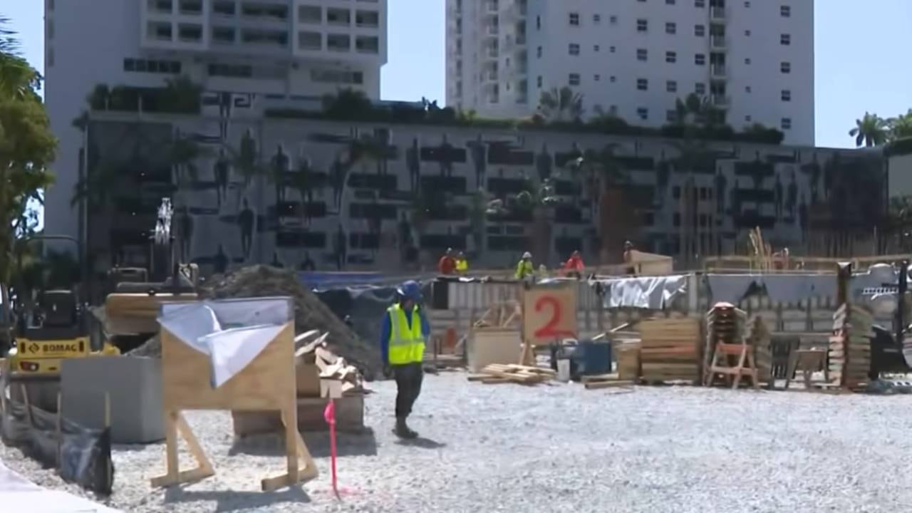 Trabajadores y maquinaria en la obra de construcción del complejo de apartamentos Plaza M, con edificios de gran altura visibles al fondo. (CBS Miami)