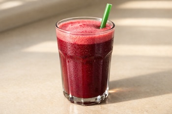 Primer plano de un vaso de batido rojo intenso con espuma rosada y gotas de condensación, con un popote verde, sobre una superficie beige bajo luz natural.